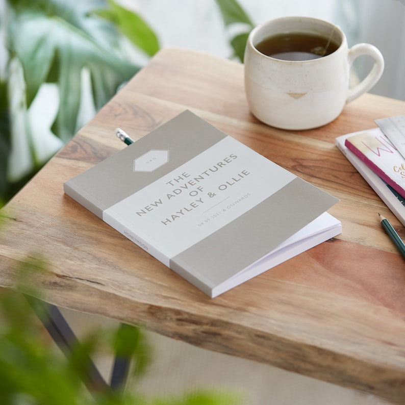 Pu&ograve; includere: Un diario grigio chiaro intitolato "The New Adventures of Hayley & Ollie" con testo bianco, aperto su un tavolo di legno. Sono presenti anche una tazza di t&egrave; e una matita. La copertina del diario include la data "04.09.2021 & onwards".