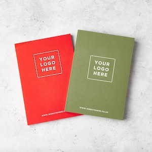 Może przedstawiać: Dwa notatniki w twardej oprawie, jeden czerwony i jeden oliwkowy, są wyświetlane na jasnoszarej powierzchni. Każdy notatnik ma biały kwadrat z tekstem "YOUR LOGO HERE" i adresem strony internetowej www.websitehere.co.uk.