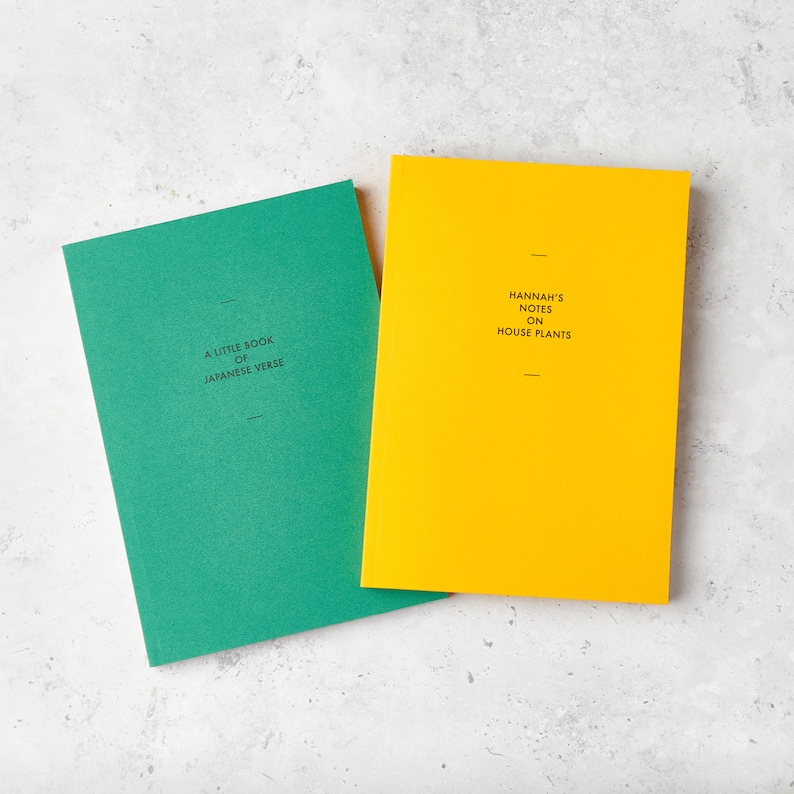 May include: Two hardcover notebooks. One is emerald green with the text "A Little Book of Japanese Verse." The other is yellow with the text "Hannah's Notes on House Plants." Both have a minimalist design with a single line above and below the text.