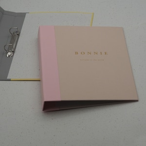 May include: A beige and light pink binder with the name "BONNIE" in gold lettering. The text "welcome to the world" is below the name. A gray binder with metal rings is visible in the background.