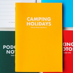 May include: A yellow notebook with the text "CAMPING HOLIDAYS" and "thomas family adventures" in white. The notebook is on a table with other notebooks.