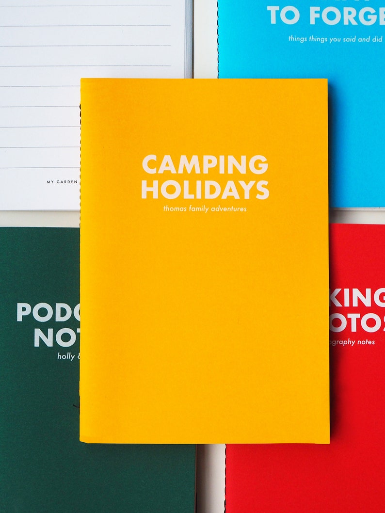 May include: A yellow notebook with the text "CAMPING HOLIDAYS" and "thomas family adventures" in white. The notebook is on a table with other notebooks.