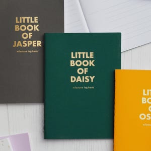 Op de afbeelding: Een verzameling notitieboekjes in verschillende kleuren, waaronder donkergrijs, groen en oranje. Elk notitieboekje heeft gouden letters met de tekst "Little Book of [Naam]" en "milestone log book".