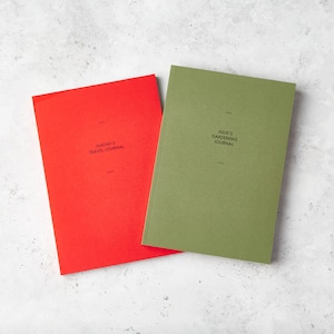 May include: Two journals, one red and one olive green, are displayed on a light gray surface. The red journal reads "NAOMI'S TRAVEL JOURNAL" and the green journal reads "JULIE'S GARDENING JOURNAL". Both journals have a minimalist design.