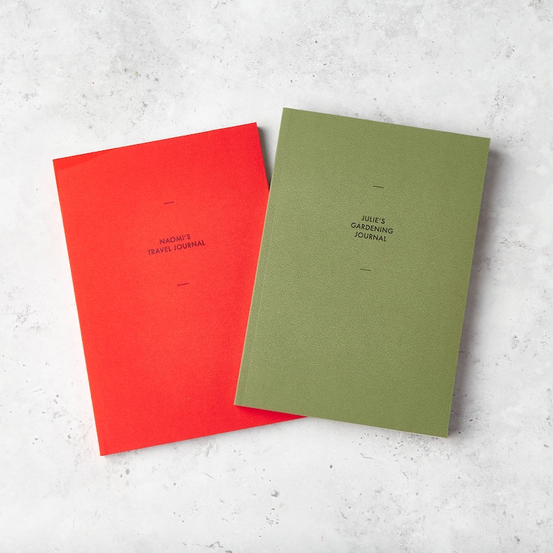 May include: Two journals, one red and one olive green, are displayed on a light gray surface. The red journal reads "NAOMI'S TRAVEL JOURNAL" and the green journal reads "JULIE'S GARDENING JOURNAL". Both journals have a minimalist design.