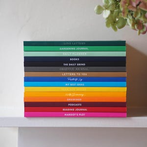 Może przedstawiać: Stos kolorowych dzienników z tytułami takimi jak "Love Letters", "Gardening Journal" i "Daily Planner". Dzienniki są w różnych kolorach, w tym zielonym, niebieskim, żółtym i różowym. Tekst jest w czcionce bezszeryfowej.