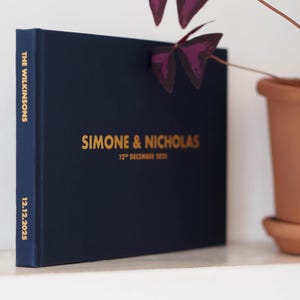 Könnte beinhalten: Ein dunkelblaues Hardcover-Buch mit goldener Folienprägung auf dem Einband und dem Buchrücken. Auf der Vorderseite steht SIMONE & NICHOLAS und 12TH DECEMBER 2025. Der Buchrücken zeigt THE WILKINSONS und 12.12.2025. Das Buch steht aufrecht.