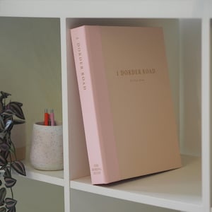 May include: A hardback book with a light pink spine and a tan cover, featuring the text 1 DORDER ROAD and THE BORING STUFF printed in gold. The book is positioned upright on a shelf next to a speckled ceramic cup containing three pens.