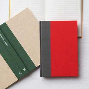Puede incluir: Dos diarios de tapa dura con cubiertas diferentes. El primer diario tiene una cubierta de lino beige con una franja verde y el texto "Morning Pages" impreso en la portada. El segundo diario tiene una cubierta roja con una franja gris.