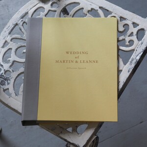 Puede incluir: Un libro de tapa dura con cubierta amarilla y lomo gris. La portada presenta el texto "WEDDING of MARTIN & LEANNE" en letras oscuras, seguido del texto más pequeño "Officiant Speech" centrado debajo de los nombres.