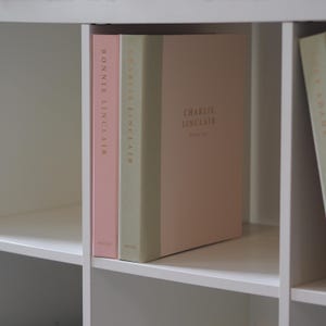 Op de afbeelding: Twee hardcover boeken met een matte kaft staan rechtop op een witte plank. Het linkerboek is lichtroze met de tekst BONNIE LINCLAIR. Het rechterboek heeft een saliegroene rug en een lichtroze kaft met de tekst CHARLIE LINCLAIR in goudkleurige letters.