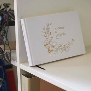 Könnte beinhalten: Ein weißes Hochzeitsgästebuch mit festem Einband, goldener Folienprägung und einem Blumenkranz-Design auf dem Cover. Der Text lautet Aaron & Louise 09.09.24, und auf dem Buchrücken steht in Gold die Aufschrift Wedding Guestbook.