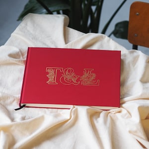 May include: A rectangular red hardcover book with a gold foil embossed design on the front cover. The design features the letters T and L intertwined with floral illustrations. A thin black ribbon bookmark extends from the bottom edge.