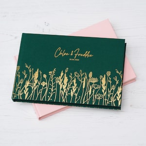 Könnte beinhalten: Ein dunkelgrünes, rechteckiges Gästebuch mit botanischen Illustrationen in Goldfolie am unteren Rand. Der Einband zeigt den Text Chloe & Freddie sowie das Datum 20.06.2025 in goldener Schrift. Ein hellrosa Buch ist darunter zu sehen.