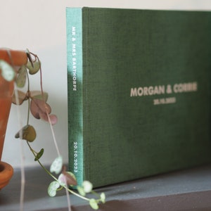 Puede incluir: Un &aacute;lbum de boda de lino verde con los nombres "Morgan & Corrie" y la fecha "20.10.2023" impresos en blanco en la portada. El lomo del &aacute;lbum dice "Mr. & Mrs. Barthorpe".