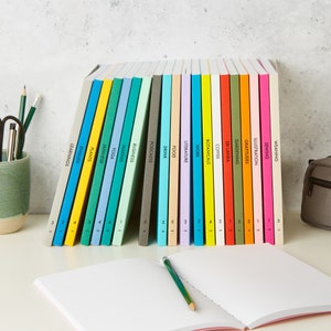 May include: A collection of colorful notebooks with labeled spines, including "Learnings," "English," "Plants," and "Japanese." A pencil rests on an open notebook in the foreground. The notebooks are arranged on a white surface.