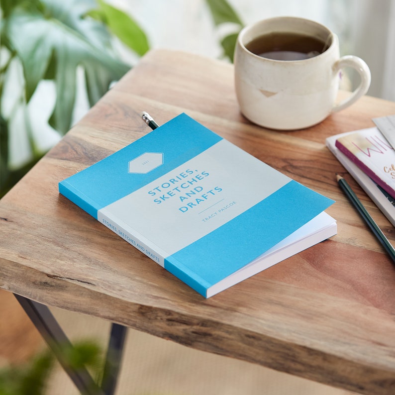 Pu&ograve; includere: Un libro intitolato "STORIES, SKETCHES AND DRAFTS" di Tracy Pascoe. Il libro ha una copertina azzurra con una sezione rettangolare bianca. Una matita &egrave; inserita nel libro. Una tazza di t&egrave; &egrave; posizionata vicino su un tavolo di legno.
