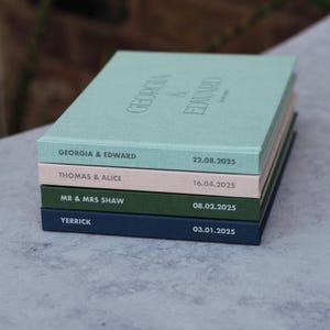 Puede incluir: Una pila de cinco cuadernos personalizados con cubiertas de diferentes colores. El cuaderno de arriba es azul claro con el texto "GEORGIA & EDWARD" y la fecha "22.08.2025". El segundo cuaderno es rosa con el texto "THOMAS & ALICE" y la fecha "16.04.2025". El tercer cuaderno es verde con el texto "MR & MRS SHAW" y la fecha "08.02.2025". El cuarto cuaderno es azul oscuro con el texto "YERRICK" y la fecha "03.01.2025".