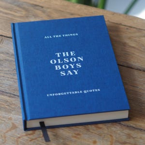 Op de afbeelding: Een gebonden boek met een diepblauwe stoffen kaft met witte tekst die ALL THE THINGS, THE OLSON BOYS SAY en UNFORGETTABLE QUOTES leest. Het boek bevat een dun zwart lint als bladwijzer dat uit de onderkant van de pagina's steekt.