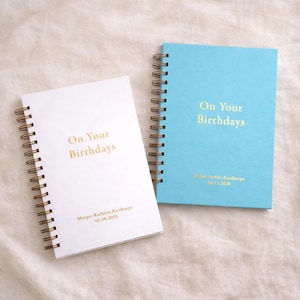 May include: Two spiral-bound notebooks, one white and one light blue, with gold lettering. The text "On Your Birthdays" is printed on the cover of each notebook. The white notebook has the name "Margot Kathryn Barthorpe" and the date "08.08.2023" printed on the cover. The blue notebook has the name "Dylan Arthur Barthorpe" and the date "16.11.2020" printed on the cover.