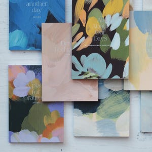 May include: A collection of six colourful journals with abstract floral designs and titles such as "Another Day", "All About Our Days", "A Place for Gratitude", and "Destination Techno".