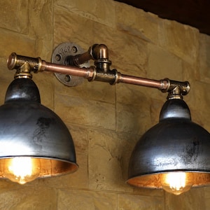 May include: A double wall light fitting constructed from copper and dark metal pipes, featuring two dome-shaped shades with a distressed grey finish. The fitting is mounted to the wall with a circular metal flange and includes two glowing bulbs.
