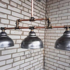 May include: Three copper pipe pendant lights with silver metal shades. The lights are hanging from a white brick wall.