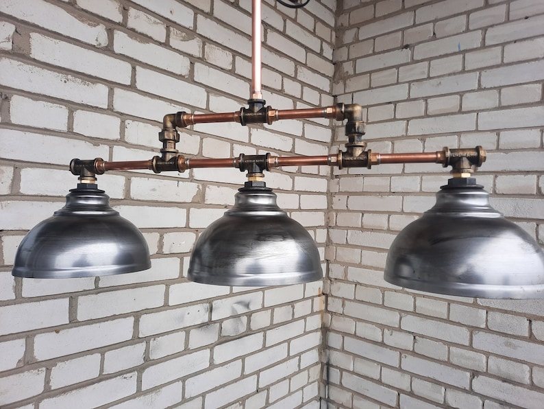 May include: Three copper pipe pendant lights with silver metal shades. The lights are hanging from a white brick wall.