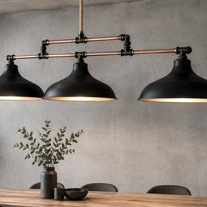 May include: A three-light pendant lamp with black shades and copper-colored pipes. The industrial-style fixture hangs above a wooden table. The shades have a gold interior, and the light is on. The fixture is a decorative element.