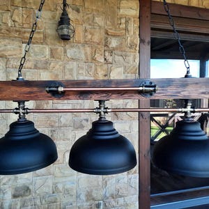 May include: A hanging light fixture featuring a horizontal dark stained wooden beam, three matte black dome-shaped metal shades, and copper piping accents. The fixture is suspended by two black metal chains and includes industrial-style pipe fittings.