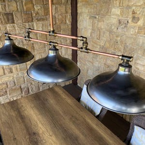 May include: Industrial-style three-light pendant lamp with a brushed metal finish. The fixture features three large, dome-shaped shades suspended from copper pipes and brass fittings. The lamp is designed to be mounted above a table.