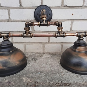 May include: Industrial-style wall sconce with two bell-shaped shades. The fixture features copper pipes, brass fittings, and a black mounting plate. The shades are black with bronze accents, creating a vintage aesthetic.