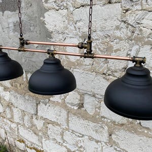 May include: A three-light pendant fixture featuring matte black dome shades suspended from a horizontal copper pipe frame with dark metal pipe fittings and connectors. The assembly is supported by two vertical metal chains attached to the top frame.
