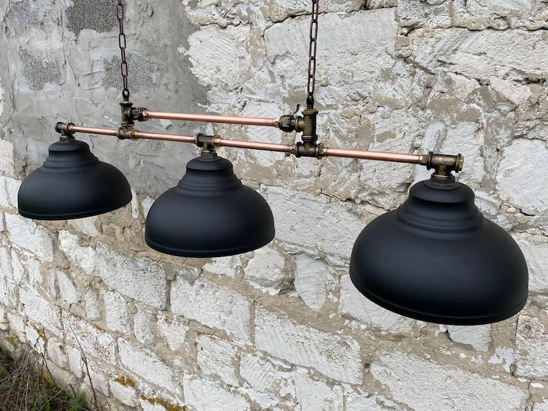 May include: A three-light pendant fixture featuring matte black dome shades suspended from a horizontal copper pipe frame with dark metal pipe fittings and connectors. The assembly is supported by two vertical metal chains attached to the top frame.