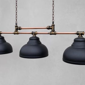 May include: A three-light pendant fixture featuring matte black dome shades suspended by a horizontal frame constructed from copper pipes and dark bronze-toned metal fittings, all hanging from two vertical chains attached to the upper pipe structure.