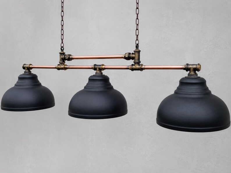 May include: A three-light pendant fixture featuring matte black dome shades suspended by a horizontal frame constructed from copper pipes and dark bronze-toned metal fittings, all hanging from two vertical chains attached to the upper pipe structure.