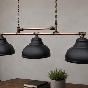 May include: A three-light pendant fixture featuring matte black dome shades suspended by copper-toned pipes and dark bronze-colored metal fittings. The structure is supported by two vertical chains, creating a linear arrangement of the three lamps.