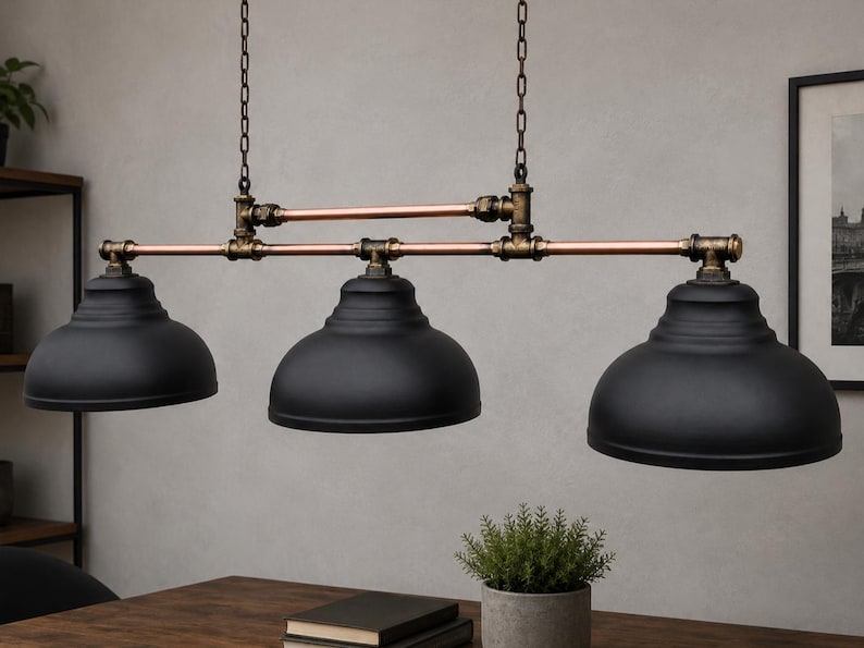 May include: A three-light pendant fixture featuring matte black dome shades suspended by copper-toned pipes and dark bronze-colored metal fittings. The structure is supported by two vertical chains, creating a linear arrangement of the three lamps.
