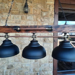 May include: A linear pendant light fixture featuring a dark stained wooden beam suspended by chains. Three matte black dome-shaped metal shades are attached to the beam with copper-colored pipes and dark metal fittings, creating a horizontal design.