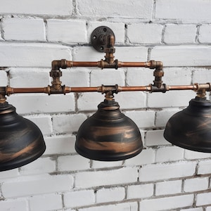 May include: A wall-mounted light fixture featuring three dome-shaped shades with a black and bronze brushed finish. The structure is composed of copper-colored metal pipes and fittings, connected to a circular mounting plate at the top center.