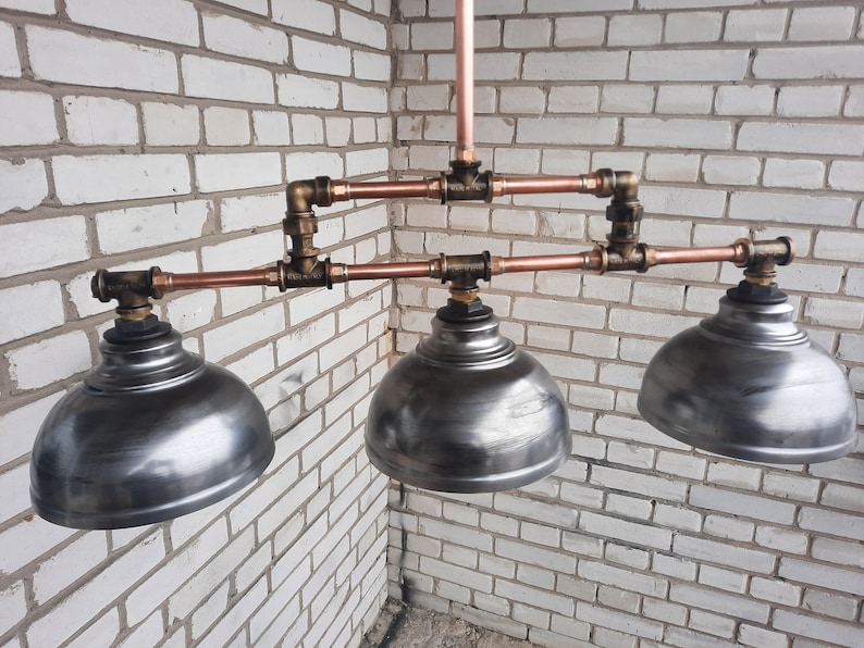 May include: A three-light industrial style pendant light fixture with copper pipes and metal shades. The shades are a brushed silver color.