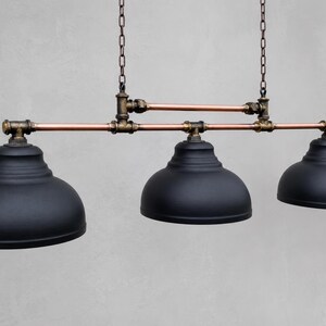 May include: A three-light pendant fixture featuring matte black dome shades suspended from a horizontal frame constructed of copper pipes and dark bronze-toned metal fittings, all hanging from two vertical chains attached to the upper pipe structure.
