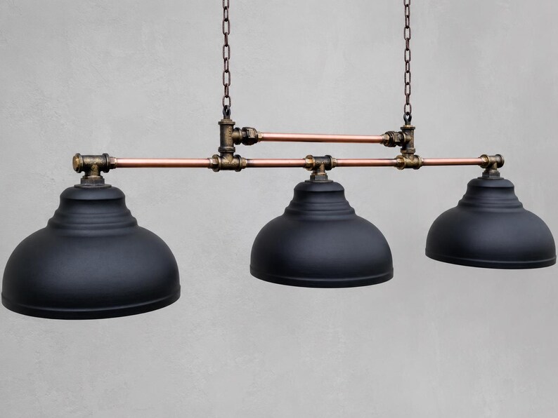 May include: A three-light pendant fixture featuring matte black dome shades suspended from a horizontal frame constructed of copper pipes and dark bronze-toned metal fittings, all hanging from two vertical chains attached to the upper pipe structure.