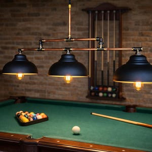 May include: A three-light, black metal pendant lamp with copper-colored pipes hangs above a green felt pool table. The lamp has three black shades with exposed Edison-style bulbs. A rack of pool cues and a triangle of pool balls are visible.
