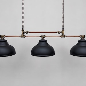 May include: A three-light pendant fixture featuring matte black dome shades suspended by two metal chains. The structure is composed of horizontal copper pipes connected by dark bronze-toned plumbing fittings, creating a linear industrial design.