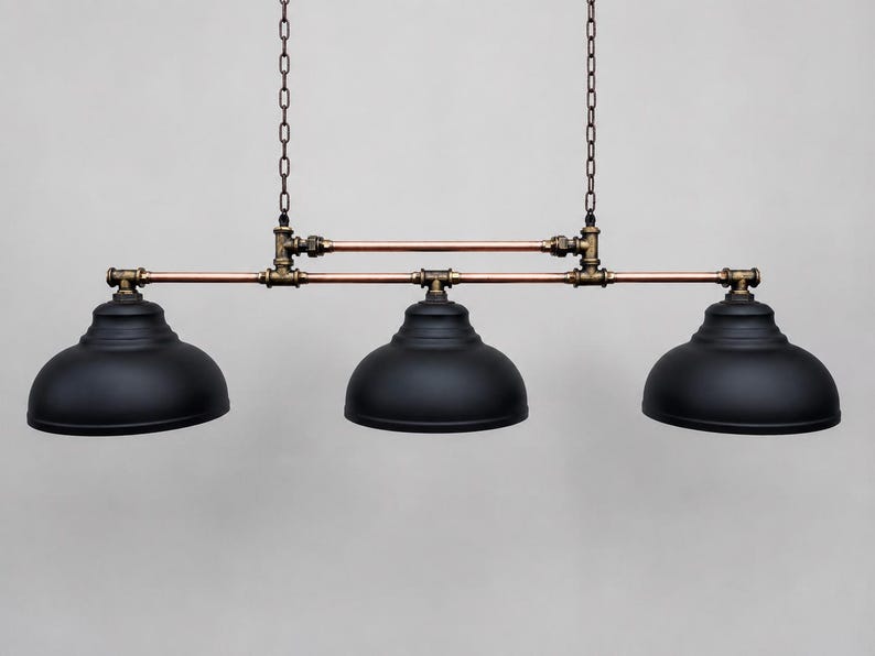 May include: A three-light pendant fixture featuring matte black dome shades suspended by two metal chains. The structure is composed of horizontal copper pipes connected by dark bronze-toned plumbing fittings, creating a linear industrial design.