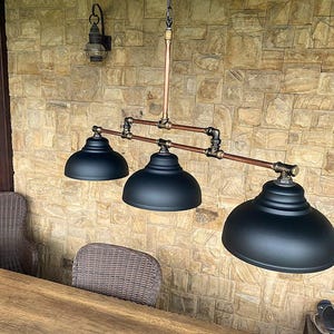 May include: A three-light pendant lamp with black dome shades, copper pipes, and brass fittings. The fixture hangs from the ceiling against a textured stone wall. The lamp is designed in an industrial style.