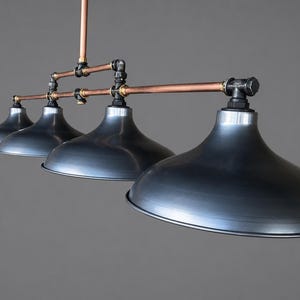 May include: An industrial pendant light with four grey metal shades. The fixture has copper pipes and black fittings, giving it a vintage look. The bell-shaped shades hang from a central support, creating a striking visual.