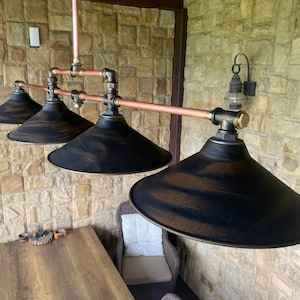 May include: A four-light pendant fixture featuring a horizontal copper pipe frame with black metal fittings and four wide, conical shades in a distressed black finish with bronze-toned highlights, suspended from a central copper pipe.