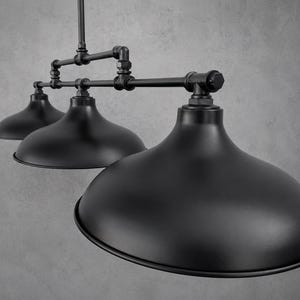 May include: A matte black triple pendant light fixture featuring three wide dome-shaped shades suspended from a central horizontal pipe structure. The assembly is constructed with industrial-style metal pipes, connectors, and fittings in a matching black finish.